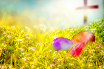 Easter background with colorful painted Easter eggs in the grass, with spring flowers and a cross in the background. Easter resurrection religious background with copy space for text.