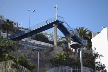 Pedestrian metal bridge in the mountains against the background of buildings and palm trees.