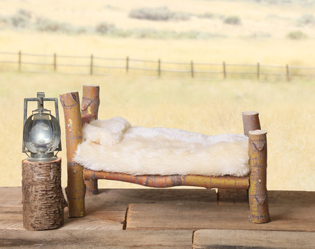 A newborn backdrop background photography prop with a rustic log bed, a railroad lantern on a nightstand, a faux cream bedspread and a yellow country meadow with a fence in the background.