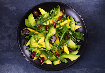 Avocado, mango, red grape and arugula salad on black plate. View from above, top studio shot