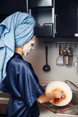 The girl washes the dirty dishes with a frying pan in a nourishing mask on the skin. Mask for...