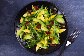 Avocado, mango, red grape and arugula salad on black plate. View from above, top studio shot