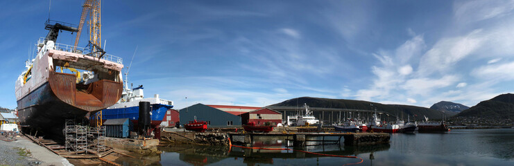 Panorama einer Werft im Hafen von Tromso, Norwegen, mit Schiff auf dem Trockendock, Ölsperre, Lagerhallen, Trawler, Fischkutter und der Tromsøbrua im Hintergrund