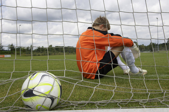 Disappointed goalkeeper sitting in goal, ball in net