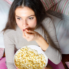 Beautiful young girl watching horror movie late at night and eating popcorn