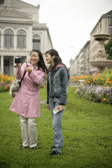 Fototapeta na wymiar Young Asian woman with a camera standing next to a younger woman in front of a park and is laughing, selective focus