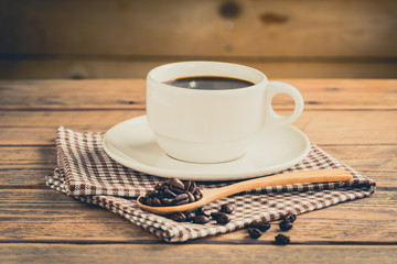 Coffee beans in spoon with a cup of coffee on wood table.