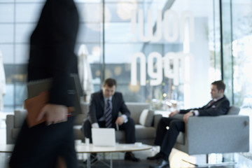 Businessmen sitting in a lobby, a businesswoman passing