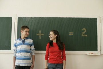 Two pupils standing in front of a blackboard, arithmetic problem in background