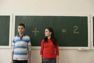 Two pupils standing in front of a blackboard, arithmetic problem in background
