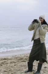 Young brownhaired Woman with woolen Hat and Rubber Boots looking through Binoculars - View - Leisure Time - Coldness - Beach