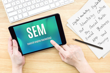 Hand using tablet with SEM (Search engine marketing ) word on screen with list of digital marketing features on paper on work table,Online business concept