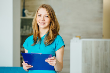 Portrait of young beautician. Young female receptionist holding blue folder and standing in nice modern hall of cosmetologycal clinic. Beauty salon interior - obrazy, fototapety, plakaty