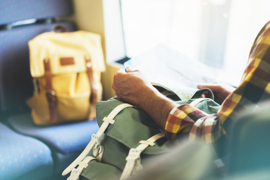 Enjoying travel. Young hipster smile man with backpack traveling by train sitting near the window holding in hand and looking map. Tourist in summer shirt planing route of railway, railroad transport
