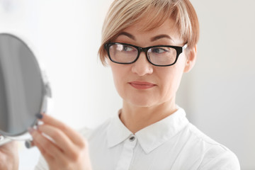 Mature woman with mirror trying on new glasses in ophthalmologist's office