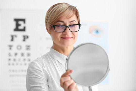 Mature woman with mirror trying on new glasses in ophthalmologist's office - Powered by Adobe