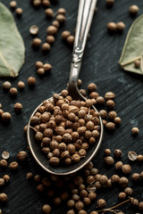 Coriander dry seeds in metal spoon on a black wooden table, vertical