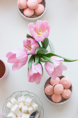 Spring background. Pink Macarons, lump sugar, a cup of tea and a bouquet of pink tulips on a white table, top view. Still life with fresh bouquet of tulips. Beautifully decorated tray.