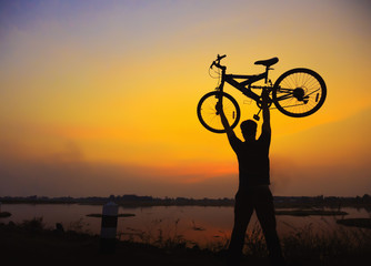 Silhouette the man stand in action lifting bicycle above his head on the meadow with sunset. Concept of arrival.