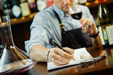 Male sommelier tasting red wine and making notes at bar counter
