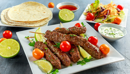 Homemade Kofta kebabs on skewers with pita, lime, vegetables, sweet chili and yogurt sauce on white plate.