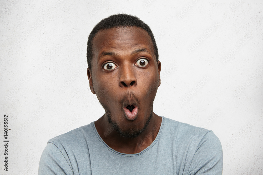 Wall mural Headshot of goofy surprised bug-eyed young dark-skinned man student wearing casual grey t-shirt staring at camera with shocked look, expressing astonishment and shock, screaming 