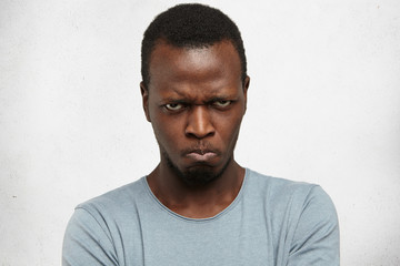 Obraz premium Studio portrait of displeased, angry, grumpy and pissed off young African American male looking and frowning at camera, pursing lips, having bad mood, feeling dissatisfied and furious with something