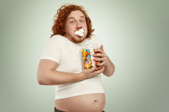 Obesity, gluttony, people and unhealthy lifestyle. Funny fat obese redhead man in white t-shirt, his belly showing up, holding jar of goodies, obsessed with food, cramming up mouth with marshmallow