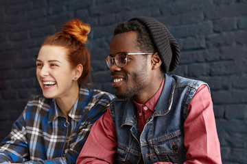 Side view of happy stylish young couple having fun indoors: handsome dark-skinned hipster wearing trendy hat and glasses relaxing at coffee shop, sitting next to his cheerful Caucasian girlfriend