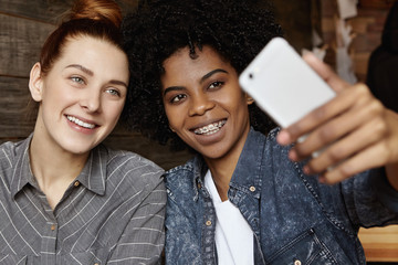 Close up shot of charming redhead lesbian with hair knot posing for selfie together with her hipster female partner wearing braces, both looking at camera of generic mobile phone and smiling happily