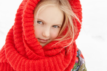 Outdoor view of cute European female child with blonde hair posing outside during short walk in city park in cold winter morning, looking at camera with faint smile, wrapped in red knitted scarf