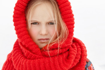 Headshot of beautiful blonde Caucasian little girl wearing red woolen scarf around head and neck, having unhappy facial expression, disliking windy cold weather while walking outside on winter day