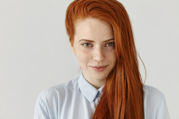 Portrait of gorgeous young European female student with freckles wearing her long ginger hair on the side, looking at camera with joyful charming smile, having good mood before going out to college