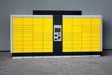 Yellow self-service parcel terminal on the background of gray wall and pavement.