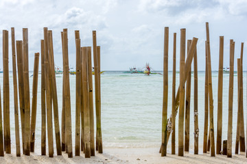 Looking through a bamboo pole barrier fence in a tropical island white sand beach out to a calm crystal clear sea with pump boats anchored in the distance with background focus.