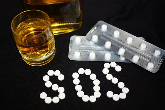 White pills on black background, which forming the word SOS, with glass and bottle of alcohol. Benzodiazepines suicide combination or addiction narcotic concept.