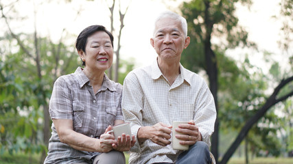 Old Asian senior couple morning date with cup of coffee in green background park