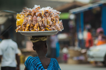 African woman carrying plantain and cassava chips on her head as she walks through a market in Cape Coast, Ghana - obrazy, fototapety, plakaty