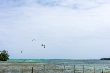 A group of kite surfs far out surfing in a calm sea viewed from a sheltered bay with the head land either side and fence poles in the water in the foreground.