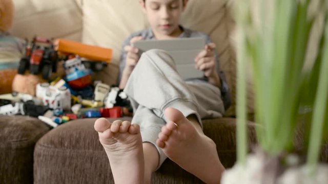 Boy plays games on the tablet, and sits on the bed beside toys