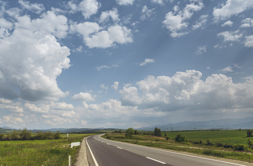 Fototapeta premium Sunny summer landscape with fluffy clouds in the blue sky and road crossing vast green plains.