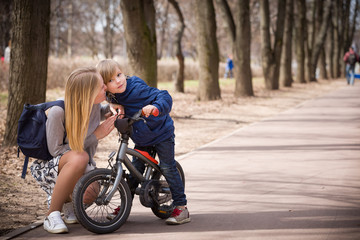 Fototapeta na wymiar Young beautiful mother talking to her son riding on a small bicycle in the park. Child on the bike on asphalt road. Children activities outdoors. Family walking in the city park.