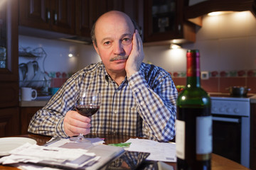 An elderly man considers a family budget and tiredly drinks red wine, trying to relax