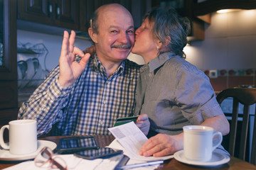 A married couple counts money and a budget. The wife kisses her husband's cheek. He shows the sign okay.