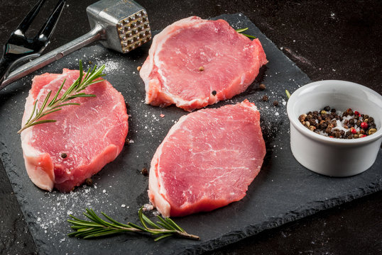 Fresh raw pork, steaks, on a cutting shale board on a black concrete table. Close view, with a hammer to beat the meat and a fork, copy space