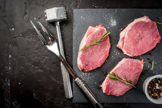 Fresh raw pork, steaks, on a cutting shale board on a black concrete table. Top view, with a hammer to beat the meat and a fork, copy space
