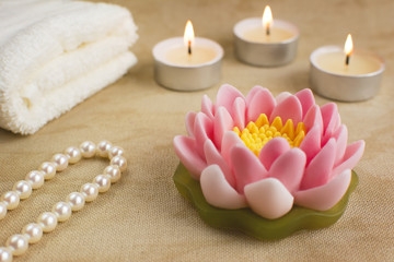Beautiful handmade soap shaped like lotus flower. Burning candles, string of pearls and towel on the background.