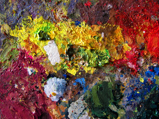 Photo of a painter's palette covered in mixed vivid paint colors. An abstract painted background that evokes a rainbow. Close-up of the brush strokes and dots texture.