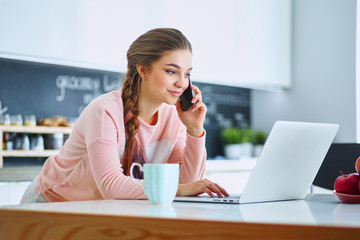 Young woman sits at the kitchen table using a laptop and talking on a cell phone