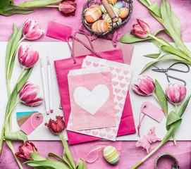 Easter decoration making. Easter workspace with eggs, tulip flowers and pink paper bags and envelope with heart, top view, copy space